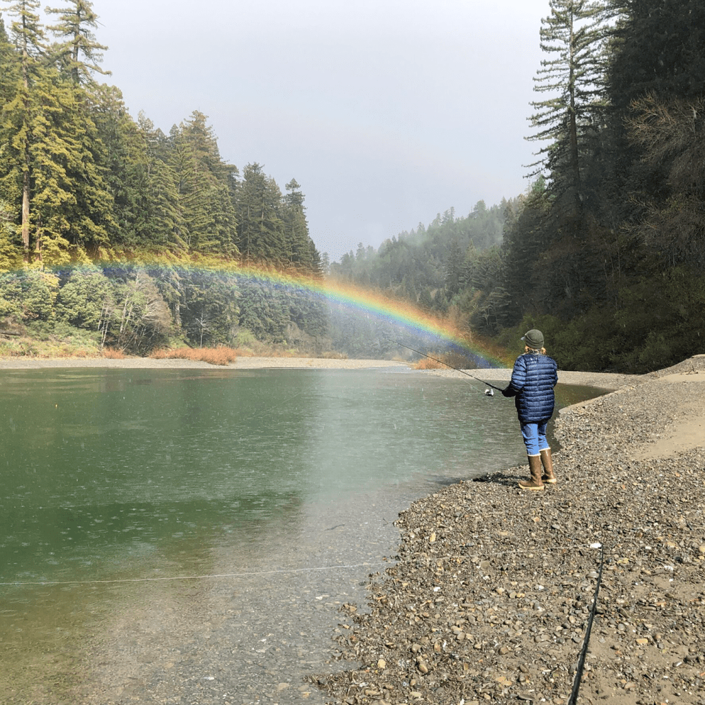 Person fishing along the coastline and rainbow just above