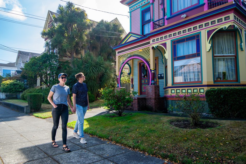 Couple holding hands walking past a rainbow colored Victorian style house