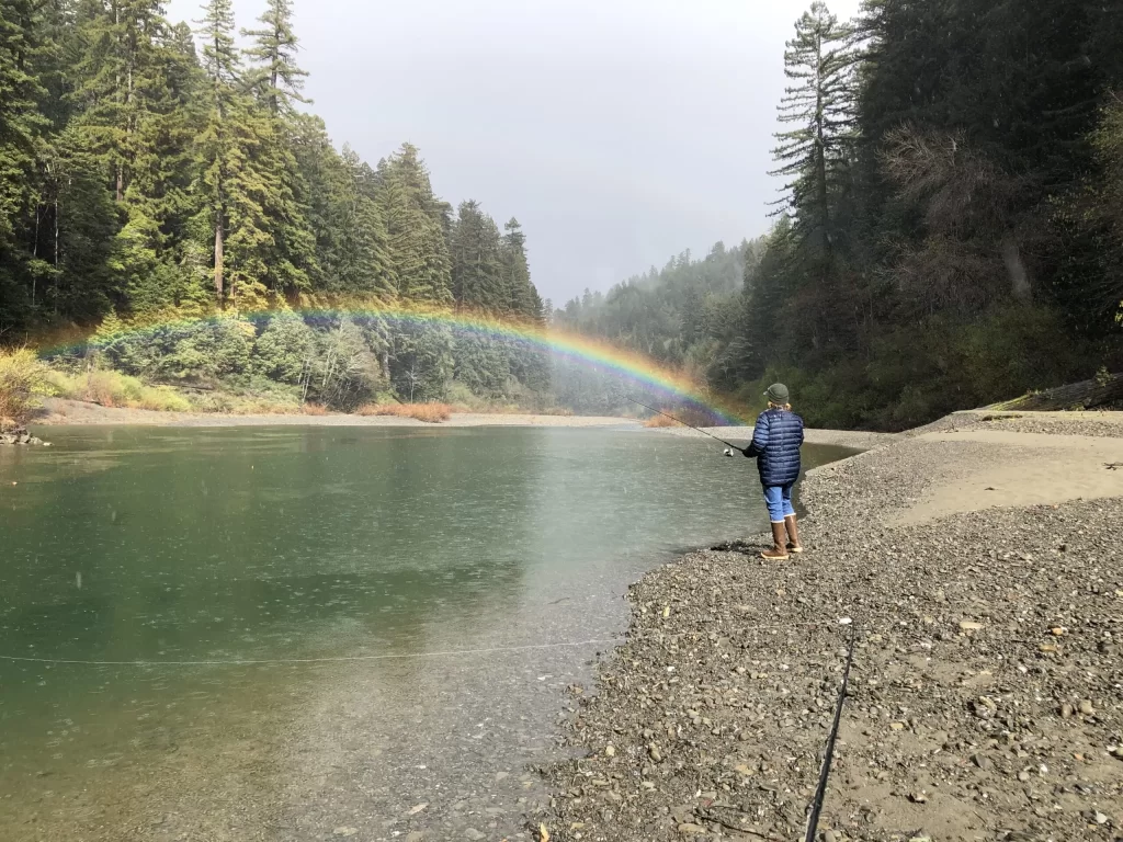 Person fishing along the coastline and rainbow just above