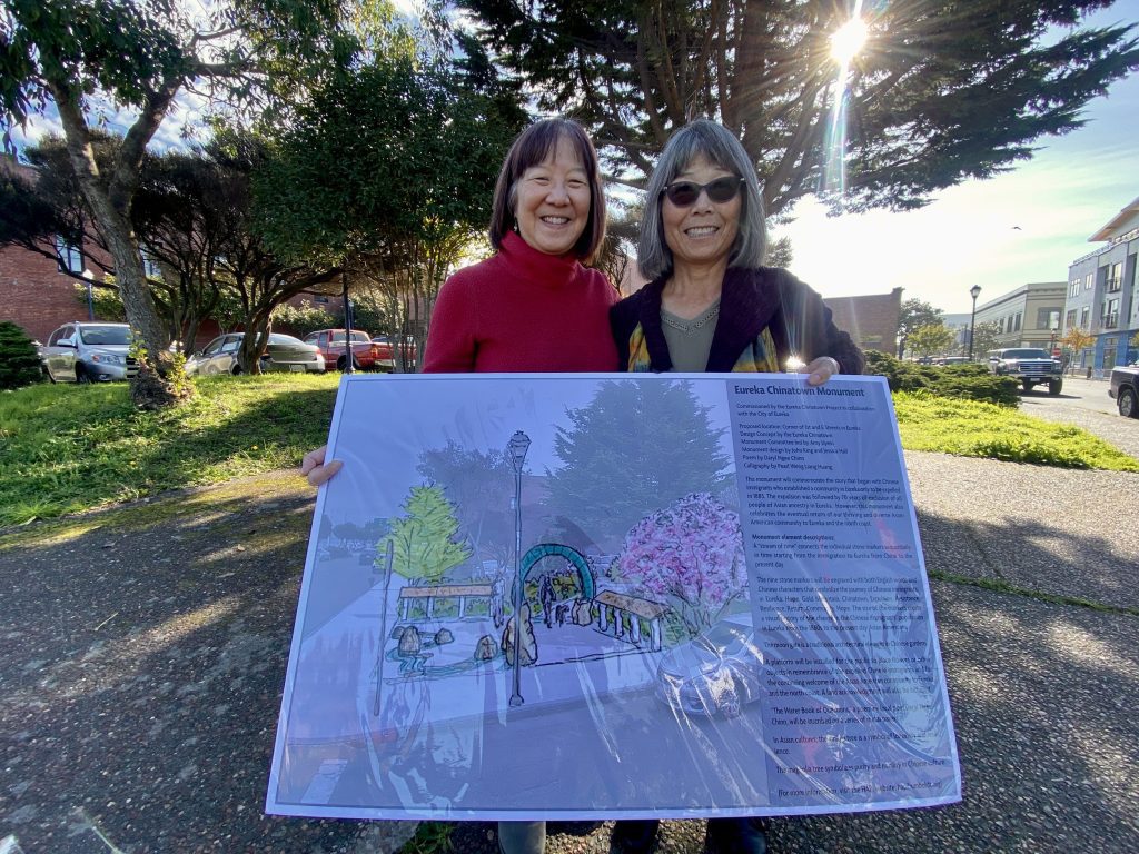 Two women standing in a park displaying a large sign with plans on it